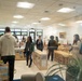 The image shows a group of adults gathered inside what appears to be a children's classroom or early education facility, likely during an open house or tour event. The room is well-lit with fluorescent ceiling lights and features child-sized furniture, educational displays, and a bookshelf with children's books. The photo is taken through a glass window or door, causing partial reflections that slightly obscure the view.