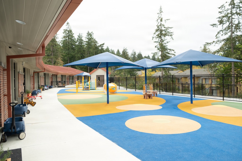 A modern, well-maintained children's playground area adjacent to what appears to be a school or daycare facility. The outdoor space features colorful rubber safety flooring in blue, yellow, and beige circular patterns, covered by large blue shade canopy structures. The area is enclosed by black metal fencing with tall evergreen trees visible in the background.