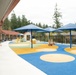 A modern, well-maintained children's playground area adjacent to what appears to be a school or daycare facility. The outdoor space features colorful rubber safety flooring in blue, yellow, and beige circular patterns, covered by large blue shade canopy structures. The area is enclosed by black metal fencing with tall evergreen trees visible in the background.