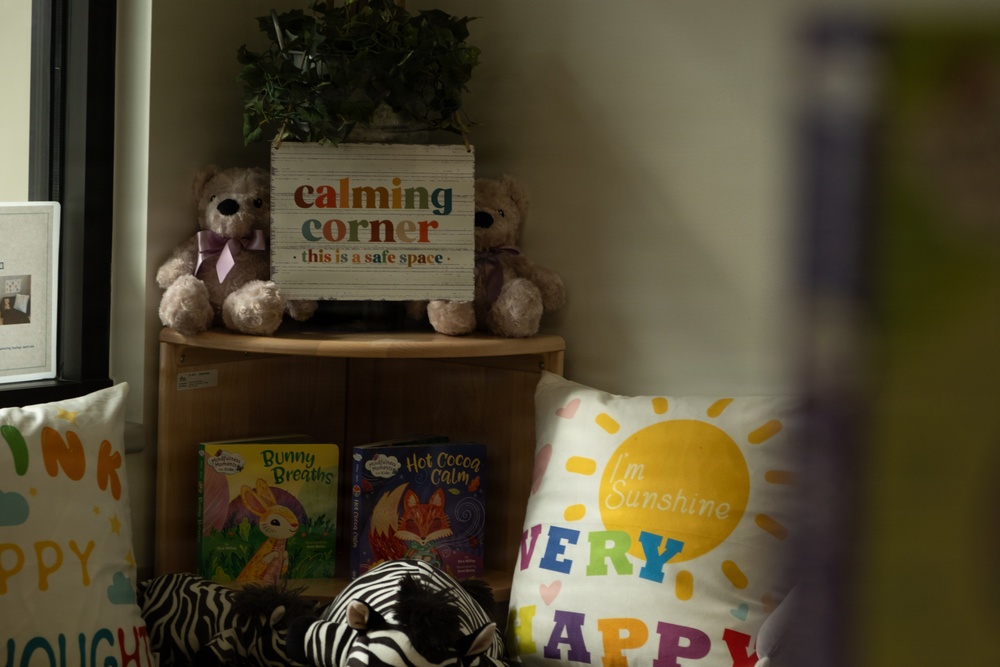 The image shows a children's 'Calming Corner' setup, likely in a classroom or therapeutic space. It features a corner shelf unit with stuffed animals, books, and decorative pillows designed to create a safe, soothing environment for children. The space includes mindfulness-themed books and cheerful decor intended to help children regulate their emotions.