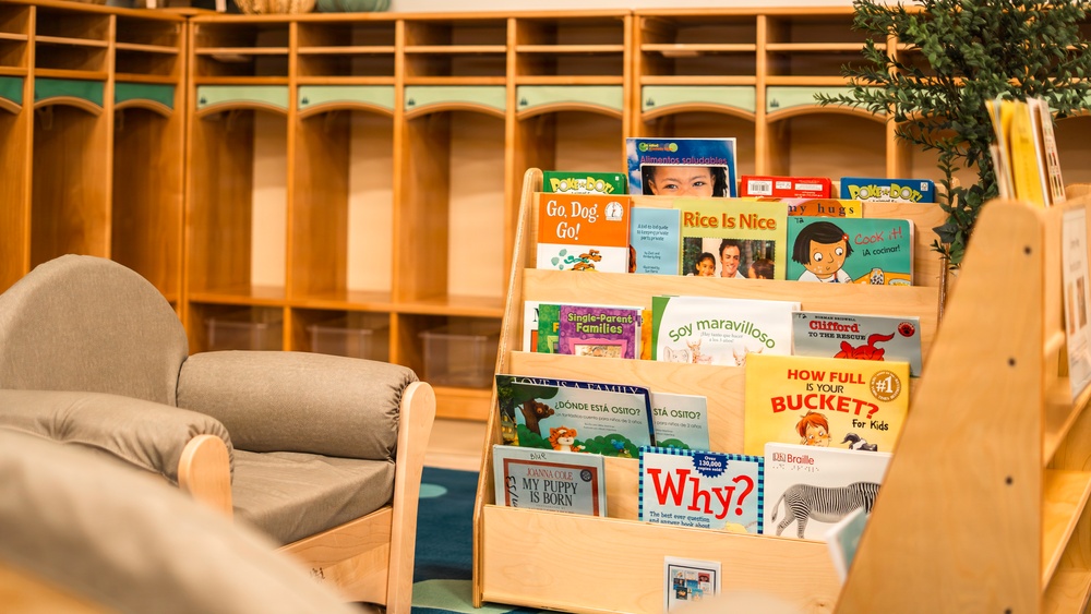 A well-organized children's reading corner in what appears to be a classroom or daycare setting. A wooden forward-facing bookshelf displays multiple colorful children's books, positioned next to a small upholstered chair sized for young children. The background features wooden cubby storage units with teal accents, typical of early childhood education environments.