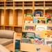 A well-organized children's reading corner in what appears to be a classroom or daycare setting. A wooden forward-facing bookshelf displays multiple colorful children's books, positioned next to a small upholstered chair sized for young children. The background features wooden cubby storage units with teal accents, typical of early childhood education environments.