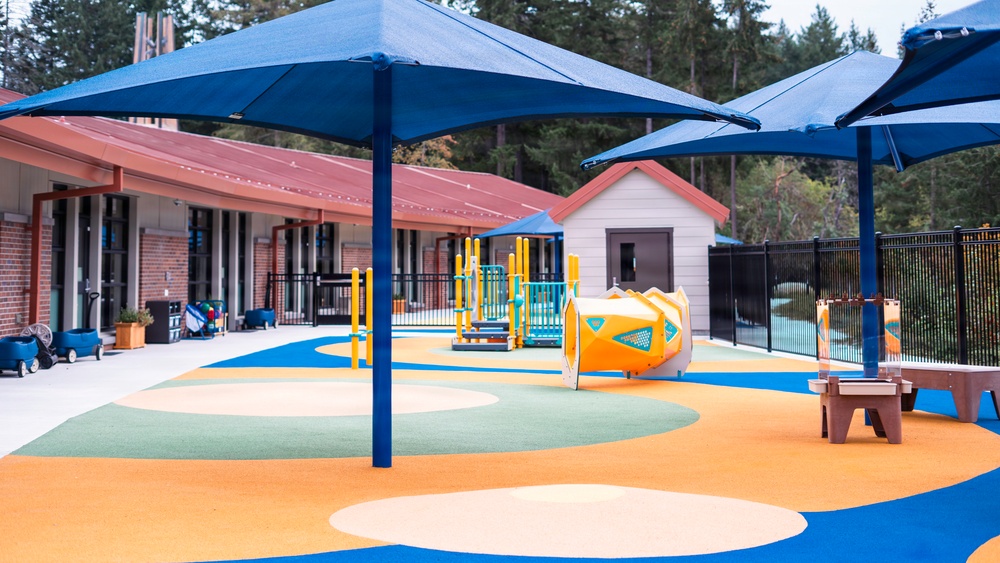 A modern, well-equipped children's playground at what appears to be a school or daycare facility. The area features colorful rubber safety surfacing in blue, orange, green, and cream swirl patterns, covered by large blue shade canopies. The facility is surrounded by tall evergreen trees and includes a brick and siding building complex.