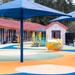 A modern, well-equipped children's playground at what appears to be a school or daycare facility. The area features colorful rubber safety surfacing in blue, orange, green, and cream swirl patterns, covered by large blue shade canopies. The facility is surrounded by tall evergreen trees and includes a brick and siding building complex.