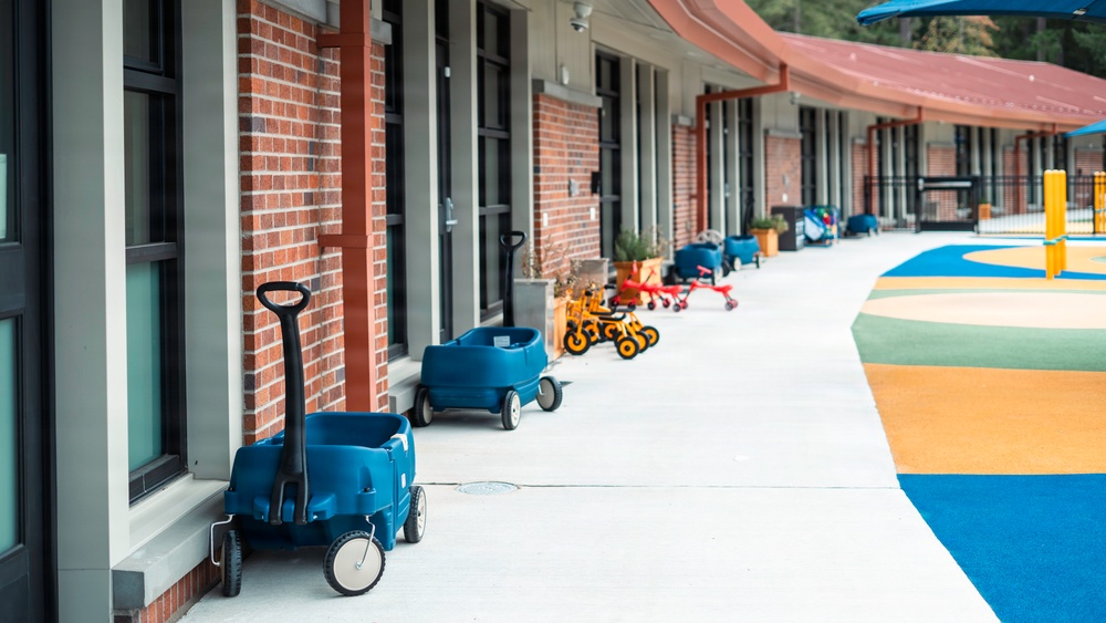 The image shows the exterior corridor of what appears to be a children's daycare or elementary school facility. Blue wagons and colorful tricycles/ride-on toys are parked along a covered walkway beside a brick building. A vibrant, multi-colored play surface is visible to the right side of the pathway.