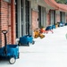 The image shows the exterior corridor of what appears to be a children's daycare or elementary school facility. Blue wagons and colorful tricycles/ride-on toys are parked along a covered walkway beside a brick building. A vibrant, multi-colored play surface is visible to the right side of the pathway.