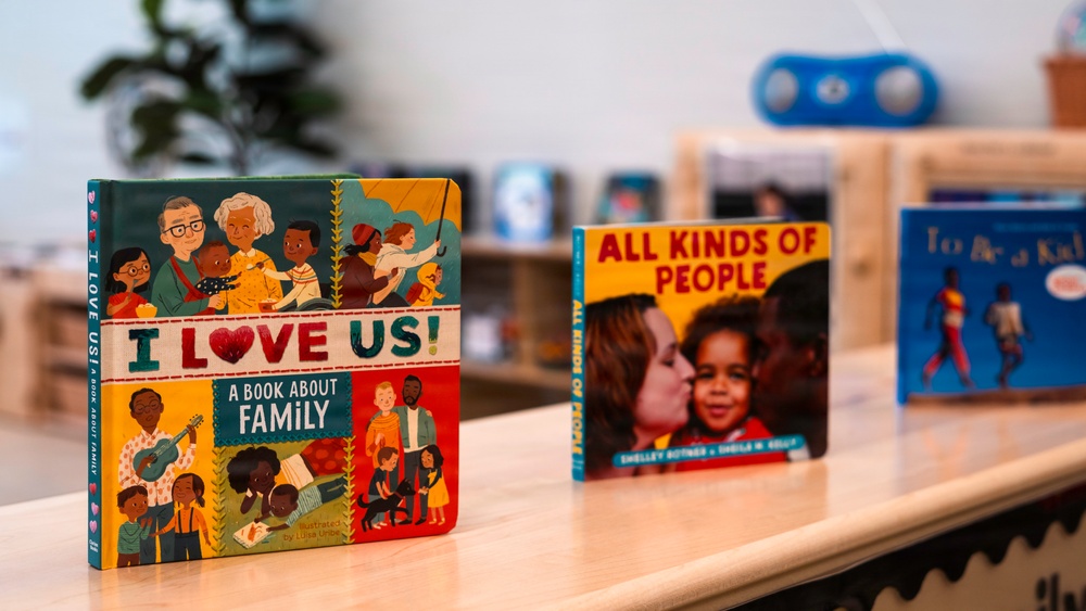 Three children's books are displayed upright on a light wooden surface, likely in a classroom or library setting. The books focus on themes of diversity, family, and inclusion. The background shows a blurred classroom environment with shelving, a plant, and what appears to be a blue speaker or toy.