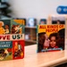 Three children's books are displayed upright on a light wooden surface, likely in a classroom or library setting. The books focus on themes of diversity, family, and inclusion. The background shows a blurred classroom environment with shelving, a plant, and what appears to be a blue speaker or toy.