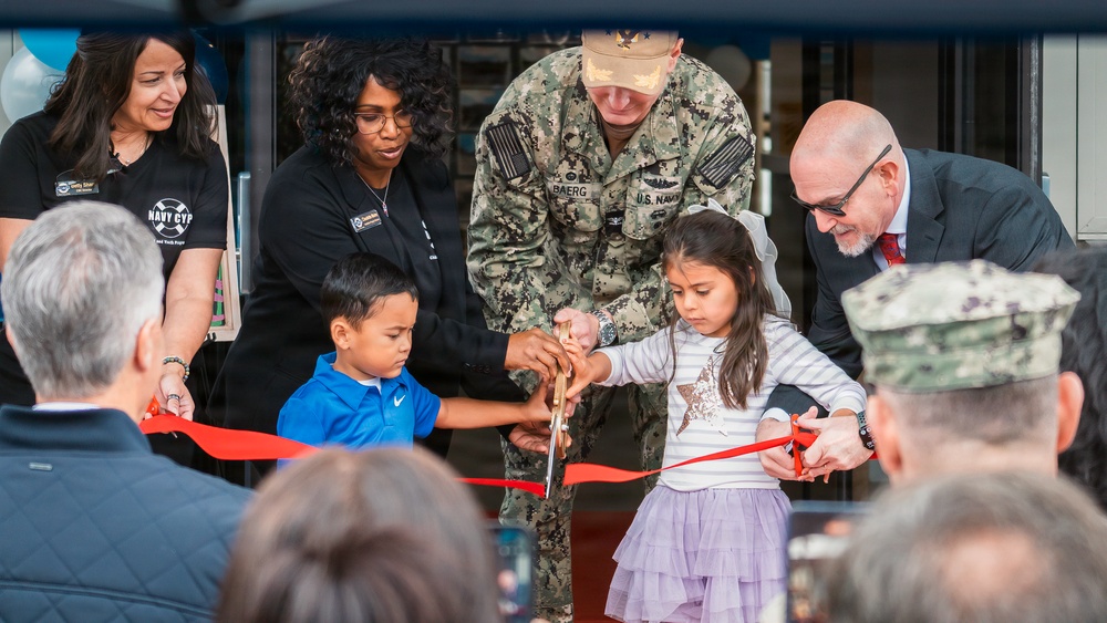 A ribbon-cutting ceremony is taking place, likely at a U.S. Navy facility, involving military personnel, civilians, and two young children. A U.S. Navy officer identified by name tape as 'BAERG' assists the children in cutting a red ribbon with large scissors. The event appears to be an official opening or dedication ceremony, with 'Navy CYP' (Child and Youth Programs) branding visible in the background.