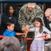 A ribbon-cutting ceremony is taking place, likely at a U.S. Navy facility, involving military personnel, civilians, and two young children. A U.S. Navy officer identified by name tape as 'BAERG' assists the children in cutting a red ribbon with large scissors. The event appears to be an official opening or dedication ceremony, with 'Navy CYP' (Child and Youth Programs) branding visible in the background.