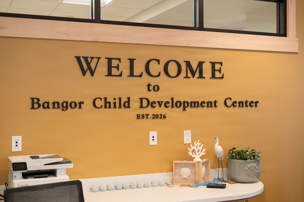 The image shows a reception or front desk area of the Bangor Child Development Center, established in 2026. The wall features large dimensional lettering reading 'WELCOME to Bangor Child Development Center EST. 2026' mounted on a warm golden-yellow wall. The desk area is decorated with coastal-themed decorative items and includes office equipment.