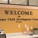The image shows a reception or front desk area of the Bangor Child Development Center, established in 2026. The wall features large dimensional lettering reading 'WELCOME to Bangor Child Development Center EST. 2026' mounted on a warm golden-yellow wall. The desk area is decorated with coastal-themed decorative items and includes office equipment.