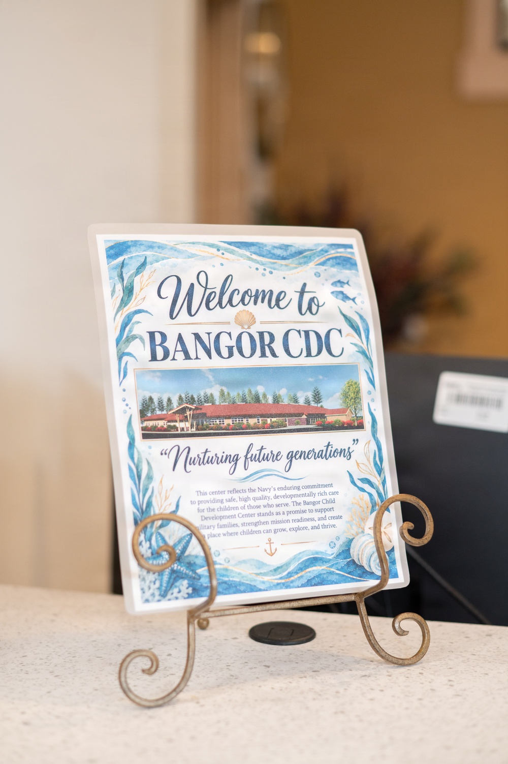 A decorative welcome sign for 'Bangor CDC' (Child Development Center) is displayed on an ornate brass easel stand on a counter or desk. The sign features an ocean/nautical theme with watercolor illustrations of seashells, starfish, seaweed, and waves. It includes a photograph of the facility building and the tagline 'Nurturing future generations.'