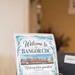 A decorative welcome sign for 'Bangor CDC' (Child Development Center) is displayed on an ornate brass easel stand on a counter or desk. The sign features an ocean/nautical theme with watercolor illustrations of seashells, starfish, seaweed, and waves. It includes a photograph of the facility building and the tagline 'Nurturing future generations.'