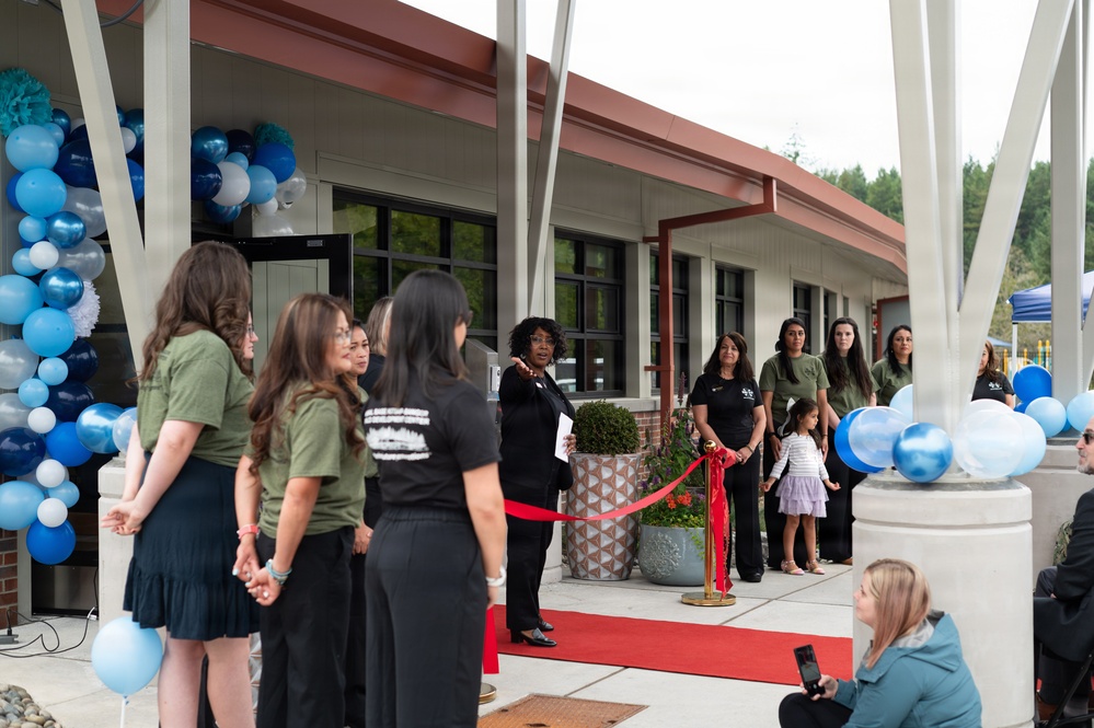 A ribbon-cutting ceremony is taking place at what appears to be a newly opened or renovated building. A woman in black attire is speaking at the center, holding papers, while a group of attendees in olive green and black shirts stand nearby. Blue, white, and navy balloon decorations frame the entrance, and a red ribbon is stretched across the walkway.