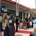 A ribbon-cutting ceremony is taking place at what appears to be a newly opened or renovated building. A woman in black attire is speaking at the center, holding papers, while a group of attendees in olive green and black shirts stand nearby. Blue, white, and navy balloon decorations frame the entrance, and a red ribbon is stretched across the walkway.