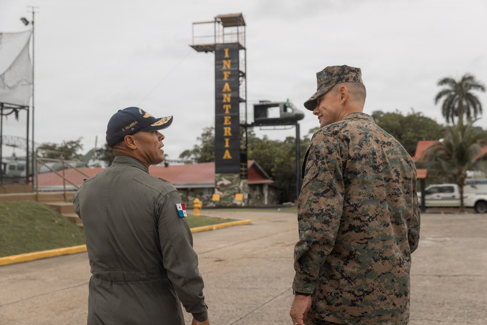Two military personnel are engaged in conversation at what appears to be an infantry training facility. One individual wears a Panamanian military flight suit with a Panama flag patch and a navy cap with the name 'E. Villarreal', while the other wears US Marine Corps MARPAT digital camouflage. They are standing in front of a tower structure marked 'INFANTERIA', suggesting a bilateral military engagement or visit.