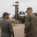 Two military personnel are engaged in conversation at what appears to be an infantry training facility. One individual wears a Panamanian military flight suit with a Panama flag patch and a navy cap with the name 'E. Villarreal', while the other wears US Marine Corps MARPAT digital camouflage. They are standing in front of a tower structure marked 'INFANTERIA', suggesting a bilateral military engagement or visit.