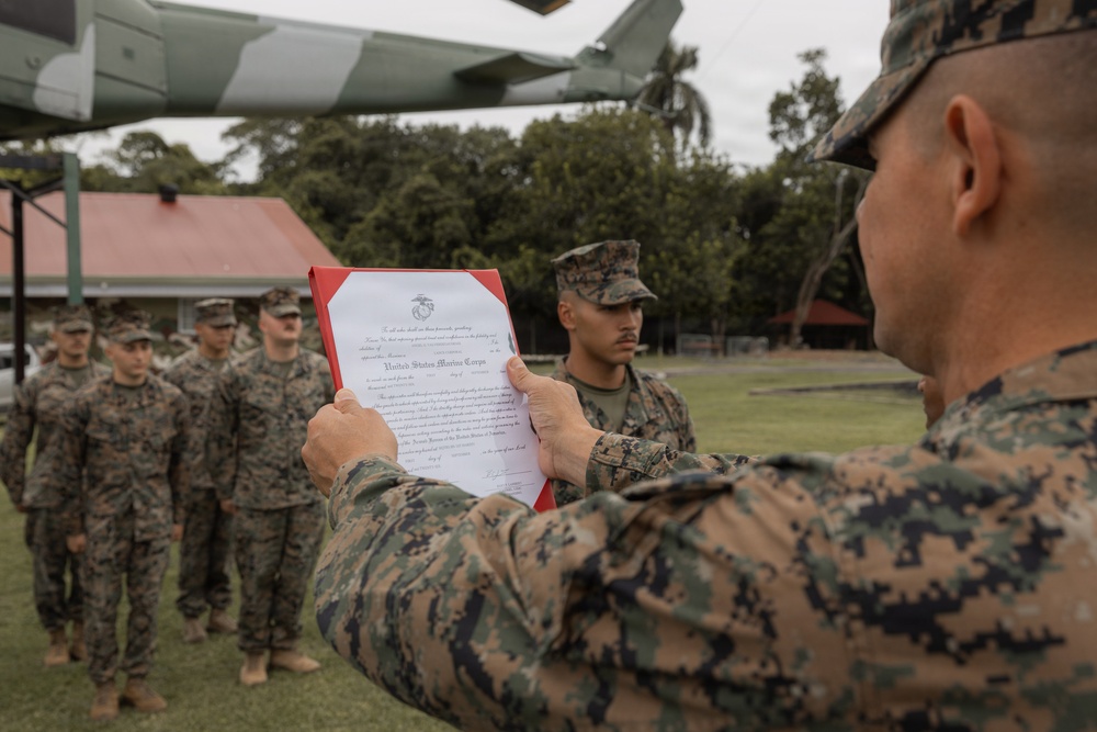 A United States Marine Corps official ceremony is taking place outdoors, where a senior Marine is reading aloud from an official USMC document held in a red folder. Several Marines in MARPAT camouflage uniforms stand at attention in formation. The setting appears to be a military installation with tropical vegetation and a partial view of an aircraft overhead.