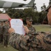 A United States Marine Corps official ceremony is taking place outdoors, where a senior Marine is reading aloud from an official USMC document held in a red folder. Several Marines in MARPAT camouflage uniforms stand at attention in formation. The setting appears to be a military installation with tropical vegetation and a partial view of an aircraft overhead.