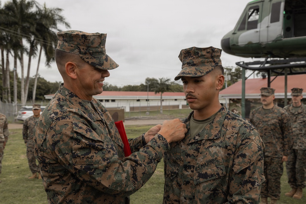 A U.S. Marines award ceremony is taking place outdoors, where a senior Marine is pinning a medal or award onto a junior Marine's uniform. The ceremony appears to be conducted on a military installation, with other Marines standing in formation in the background. A vintage military helicopter is visible on display behind the participants.