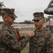A U.S. Marines award ceremony is taking place outdoors, where a senior Marine is pinning a medal or award onto a junior Marine's uniform. The ceremony appears to be conducted on a military installation, with other Marines standing in formation in the background. A vintage military helicopter is visible on display behind the participants.
