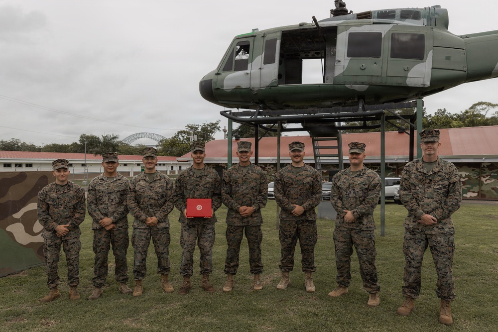 A group of eight military personnel in camouflage uniforms pose for a formal group photograph in front of a static display helicopter mounted on a metal frame. One individual in the center holds a red box or case, suggesting a ceremony or award presentation. A bridge structure is visible in the background, along with military buildings and a camouflage-painted wall.