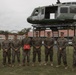 A group of eight military personnel in camouflage uniforms pose for a formal group photograph in front of a static display helicopter mounted on a metal frame. One individual in the center holds a red box or case, suggesting a ceremony or award presentation. A bridge structure is visible in the background, along with military buildings and a camouflage-painted wall.