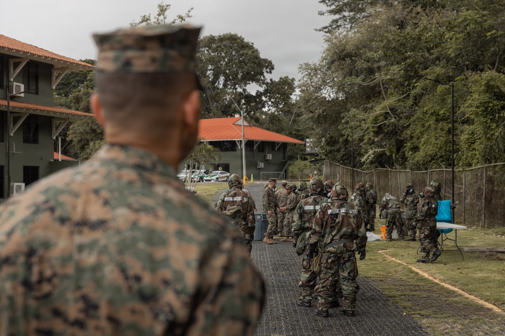 A military training exercise is taking place outdoors at what appears to be a tropical or subtropical installation, with soldiers in camouflage uniforms gathered in a formation or briefing area. A U.S. Marine in MARPAT digital camouflage is observed from behind in the foreground, watching a group of soldiers — some wearing gas masks or CBRN protective gear — assembled on a matted pathway. The setting includes green buildings with orange-tiled roofs and dense tropical vegetation.