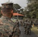 A military training exercise is taking place outdoors at what appears to be a tropical or subtropical installation, with soldiers in camouflage uniforms gathered in a formation or briefing area. A U.S. Marine in MARPAT digital camouflage is observed from behind in the foreground, watching a group of soldiers — some wearing gas masks or CBRN protective gear — assembled on a matted pathway. The setting includes green buildings with orange-tiled roofs and dense tropical vegetation.