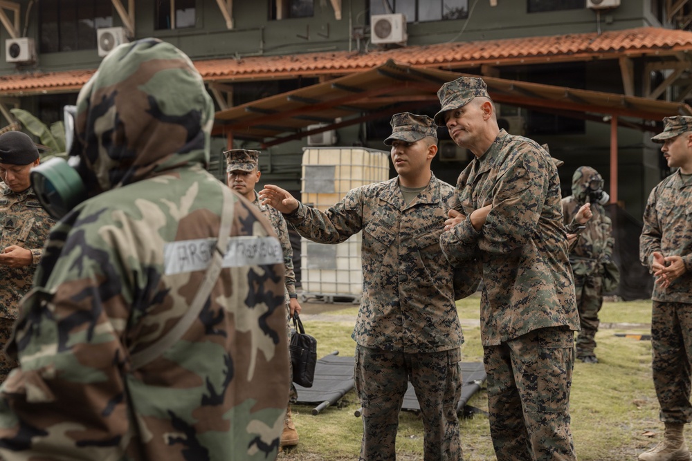 A group of military personnel in camouflage uniforms are gathered outdoors near a building, appearing to be engaged in a briefing or training exercise. One soldier in US Marine Corps MARPAT digital camouflage is gesturing and appears to be instructing or explaining something to others. Some personnel in the background are wearing gas masks, suggesting a CBRN (Chemical, Biological, Radiological, Nuclear) training scenario.