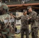 A group of military personnel in camouflage uniforms are gathered outdoors near a building, appearing to be engaged in a briefing or training exercise. One soldier in US Marine Corps MARPAT digital camouflage is gesturing and appears to be instructing or explaining something to others. Some personnel in the background are wearing gas masks, suggesting a CBRN (Chemical, Biological, Radiological, Nuclear) training scenario.