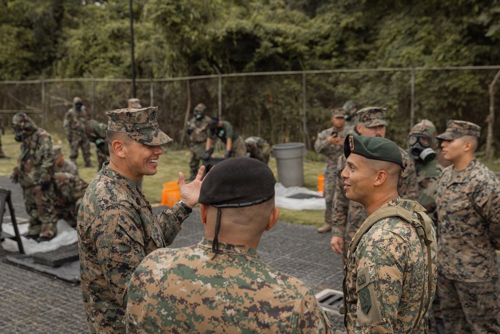 A group of military personnel from what appear to be two different nations are engaged in conversation during a joint training exercise. Some soldiers in the background are wearing gas masks, suggesting a chemical, biological, radiological, or nuclear (CBRN) training scenario. The interaction between the uniformed individuals in the foreground suggests a briefing or debriefing between officers.