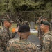 A group of military personnel from what appear to be two different nations are engaged in conversation during a joint training exercise. Some soldiers in the background are wearing gas masks, suggesting a chemical, biological, radiological, or nuclear (CBRN) training scenario. The interaction between the uniformed individuals in the foreground suggests a briefing or debriefing between officers.