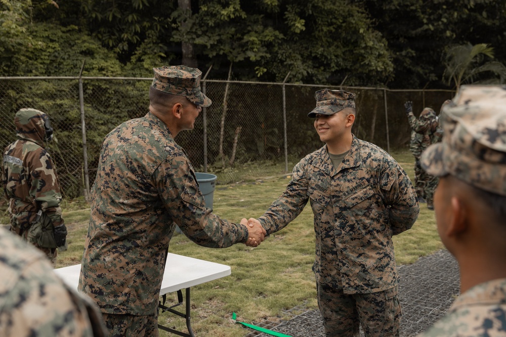 Two U.S. Marines in MARPAT woodland camouflage uniforms are shaking hands in what appears to be an outdoor ceremony or recognition event. A white folding table is visible nearby, suggesting a formal presentation setting. Additional personnel in mixed camouflage uniforms are visible in the background near a chain-link fence.