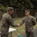 Two U.S. Marines in MARPAT woodland camouflage uniforms are shaking hands in what appears to be an outdoor ceremony or recognition event. A white folding table is visible nearby, suggesting a formal presentation setting. Additional personnel in mixed camouflage uniforms are visible in the background near a chain-link fence.