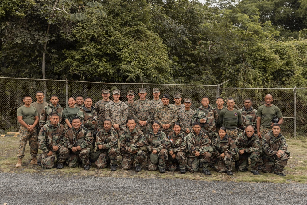 A group of approximately 30 military personnel pose for a formal group photograph outdoors. The group appears to include members wearing at least two distinct camouflage uniform patterns, suggesting a multinational or joint military exercise. They are arranged in two rows — a standing back row and a kneeling front row — in front of a chain-link fence with dense tropical vegetation behind them.