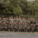 A group of approximately 30 military personnel pose for a formal group photograph outdoors. The group appears to include members wearing at least two distinct camouflage uniform patterns, suggesting a multinational or joint military exercise. They are arranged in two rows — a standing back row and a kneeling front row — in front of a chain-link fence with dense tropical vegetation behind them.