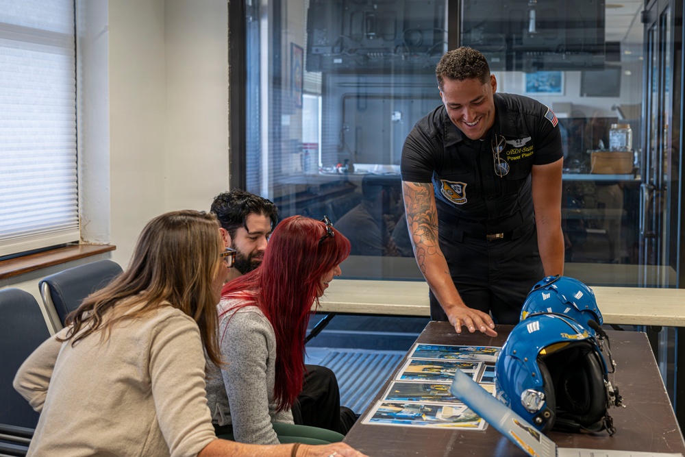 A uniformed military personnel member, wearing a black flight suit with patches including a US flag and what appears to be a Blue Angels or similar aviation team insignia, is presenting photographs or printed materials to three civilians seated at a table. A blue flight helmet is visible on the table alongside the printed materials. The setting appears to be an indoor briefing or meeting room.