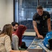 A uniformed military personnel member, wearing a black flight suit with patches including a US flag and what appears to be a Blue Angels or similar aviation team insignia, is presenting photographs or printed materials to three civilians seated at a table. A blue flight helmet is visible on the table alongside the printed materials. The setting appears to be an indoor briefing or meeting room.