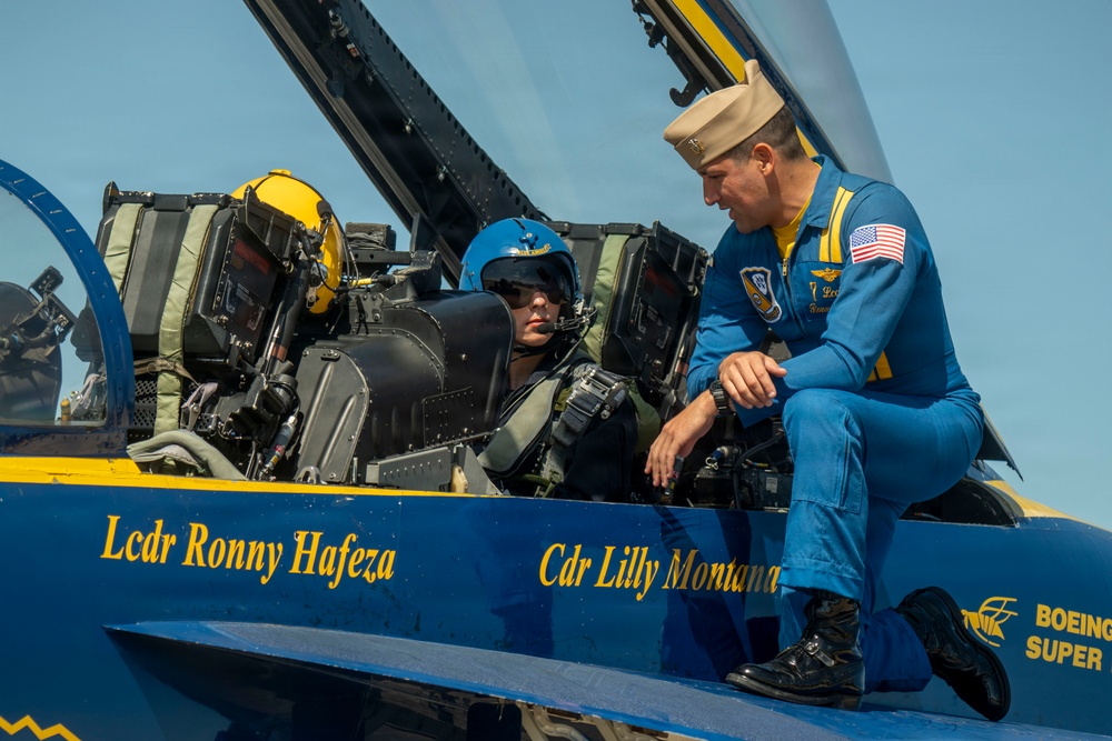 A Blue Angels U.S. Navy flight demonstration squadron aircraft is shown on the ground with two personnel. A pilot wearing a blue helmet is seated in the cockpit, while a Blue Angels officer in a blue flight suit and white cover (cap) crouches on the fuselage beside the cockpit. The aircraft fuselage displays the names 'Lcdr Ronny Hafeza' and 'Cdr Lilly Montana' in gold lettering, along with a Boeing Super Hornet logo.