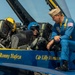 A Blue Angels U.S. Navy flight demonstration squadron aircraft is shown on the ground with two personnel. A pilot wearing a blue helmet is seated in the cockpit, while a Blue Angels officer in a blue flight suit and white cover (cap) crouches on the fuselage beside the cockpit. The aircraft fuselage displays the names 'Lcdr Ronny Hafeza' and 'Cdr Lilly Montana' in gold lettering, along with a Boeing Super Hornet logo.