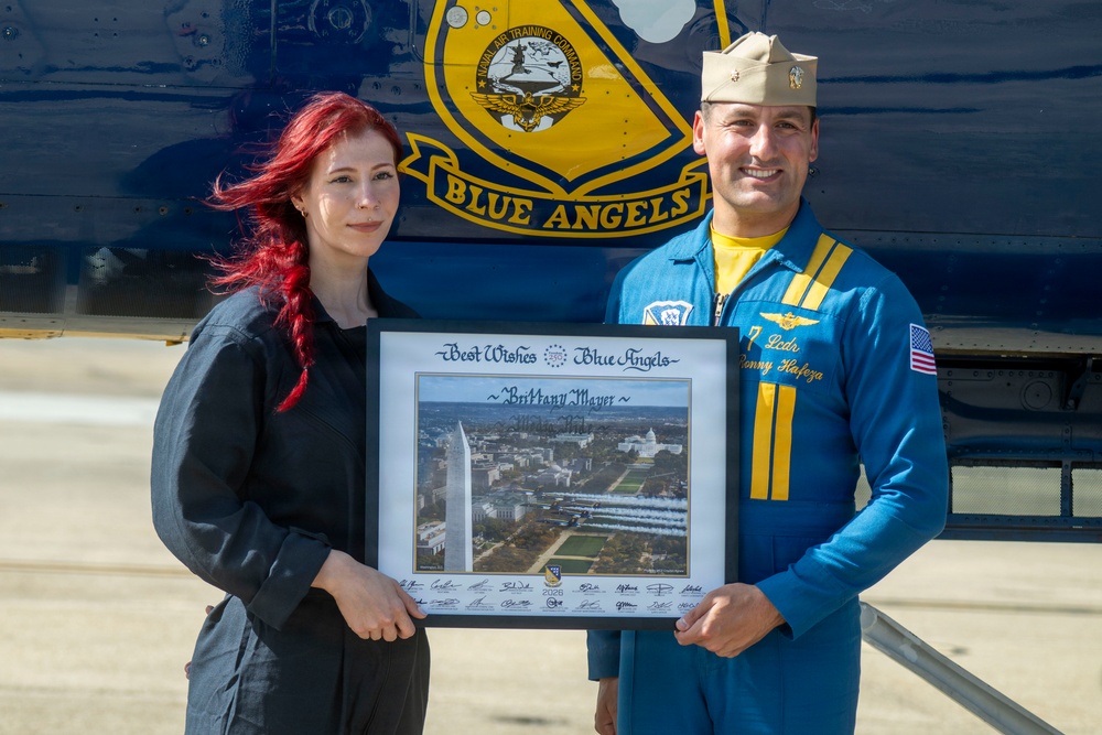 A woman with red hair and a Blue Angels pilot in a blue flight suit are posing together in front of a Blue Angels aircraft. They are jointly holding a framed commemorative photo with the inscription 'Best Wishes Blue Angels' and the name 'Brittany Mayer,' featuring an aerial view of Washington D.C. with Blue Angels jets flying over the National Mall. The pilot's name tag reads 'T. Pedr... Ronny Hafeza.'
