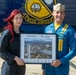 A woman with red hair and a Blue Angels pilot in a blue flight suit are posing together in front of a Blue Angels aircraft. They are jointly holding a framed commemorative photo with the inscription 'Best Wishes Blue Angels' and the name 'Brittany Mayer,' featuring an aerial view of Washington D.C. with Blue Angels jets flying over the National Mall. The pilot's name tag reads 'T. Pedr... Ronny Hafeza.'