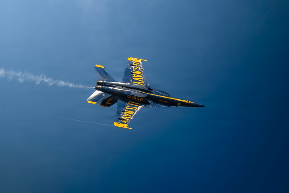 A U.S. Navy Blue Angels F/A-18 Hornet jet is captured in flight from an overhead or near-overhead angle, appearing to be inverted or in a banking maneuver. The aircraft displays the distinctive blue and gold color scheme with 'U.S. NAVY' lettering visible on the wings and fuselage. A white smoke trail is visible streaming from the rear of the aircraft against a deep blue sky.
