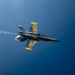 A U.S. Navy Blue Angels F/A-18 Hornet jet is captured in flight from an overhead or near-overhead angle, appearing to be inverted or in a banking maneuver. The aircraft displays the distinctive blue and gold color scheme with 'U.S. NAVY' lettering visible on the wings and fuselage. A white smoke trail is visible streaming from the rear of the aircraft against a deep blue sky.