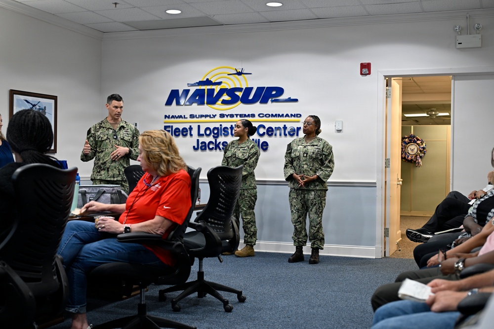 A formal military briefing or ceremony is taking place at NAVSUP Fleet Logistics Center Jacksonville, part of the Naval Supply Systems Command. Three uniformed Navy personnel, including one male and two female sailors in camouflage uniforms, are standing and addressing a seated audience of mixed military and civilian attendees. The setting is an official conference or reception room with the NAVSUP logo prominently displayed on the wall.
