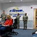 A formal military briefing or ceremony is taking place at NAVSUP Fleet Logistics Center Jacksonville, part of the Naval Supply Systems Command. Three uniformed Navy personnel, including one male and two female sailors in camouflage uniforms, are standing and addressing a seated audience of mixed military and civilian attendees. The setting is an official conference or reception room with the NAVSUP logo prominently displayed on the wall.