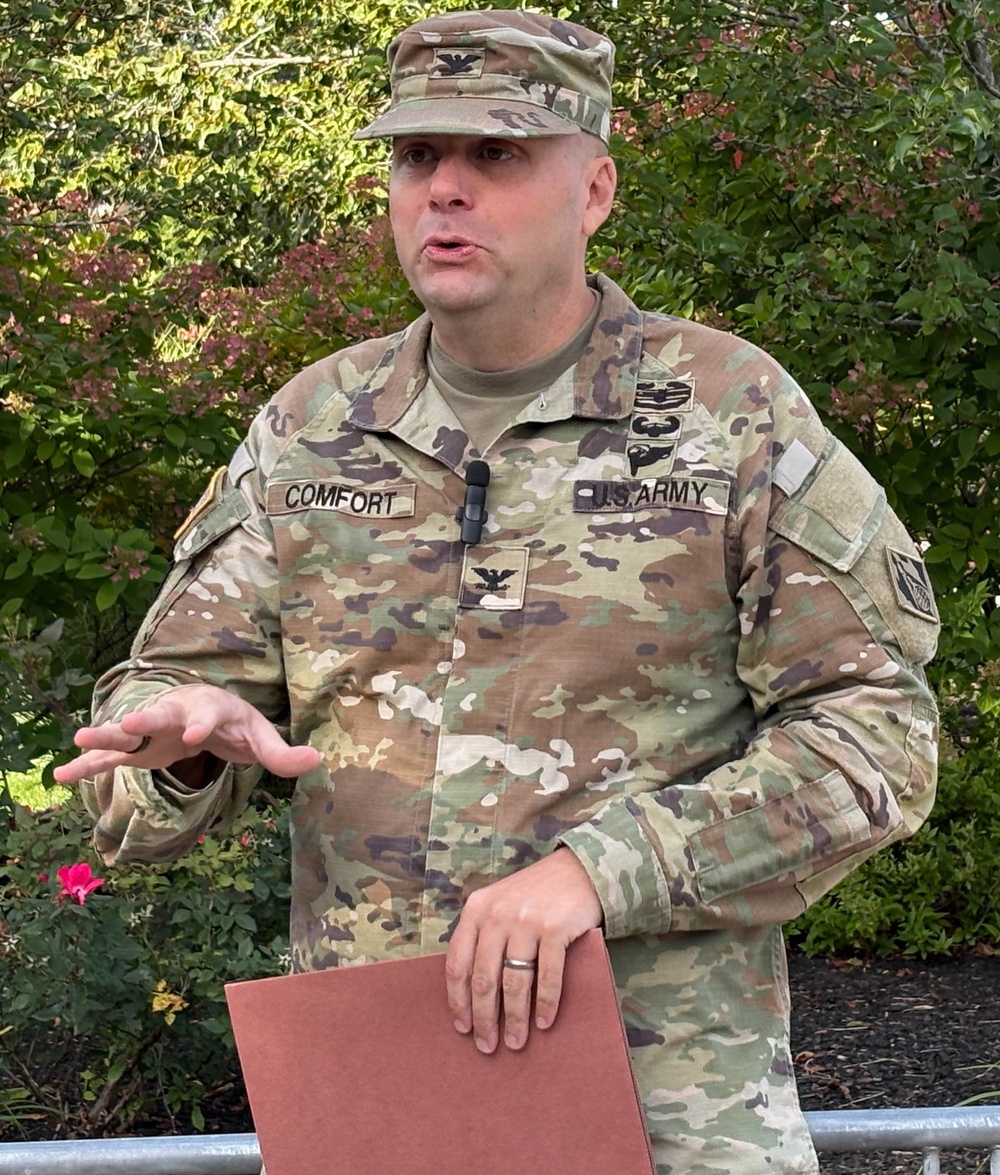 A U.S. Army Colonel named Comfort is speaking outdoors, gesturing with one hand while holding a folder in the other. He is wearing the Operational Camouflage Pattern (OCP) uniform with a Colonel's eagle insignia visible on both his cap and chest. A lapel microphone is clipped to his uniform, suggesting a formal address or press briefing.