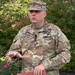 A U.S. Army Colonel named Comfort is speaking outdoors, gesturing with one hand while holding a folder in the other. He is wearing the Operational Camouflage Pattern (OCP) uniform with a Colonel's eagle insignia visible on both his cap and chest. A lapel microphone is clipped to his uniform, suggesting a formal address or press briefing.